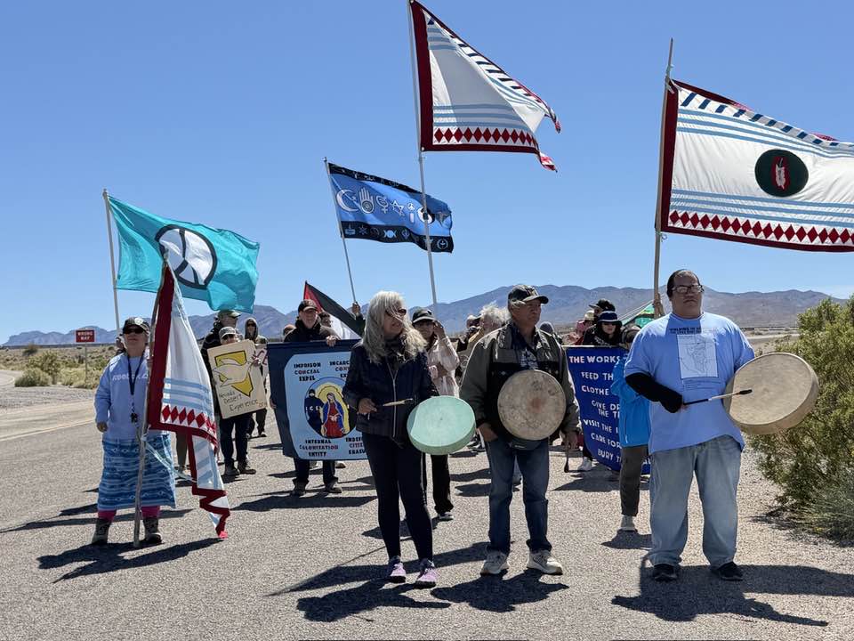 A line of marchers carry Indigenous flags, drums, and banners along a desert highway under a clear blue sky, mountains behind; a peace-symbol flag is visible.